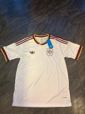 Vit Spanien fotbollströja Adidas VM 2026 - Säljer en vit fotbollströja från Adidas med Spaniens landslagsemblem på bröstet. Tröjan har vinröda detaljer, tre ränder på axlarna och guldfärgade kanter vid ärmar och hals. Tillverkad i ett lätt och svalt material, perfekt för träning eller match.