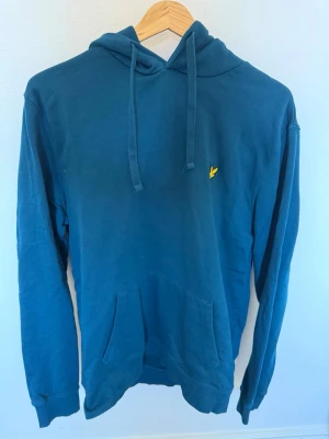 Blå hoodie med gul detalj - Snygg blå hoodie med huva och dragsnören samt en gul broderad logga på bröstet. Hoodien har en stor magficka och långa ärmar. Perfekt för en chill och avslappnad stil. Materialet känns mjukt och bekvämt, troligen bomull eller bomullsblandning.