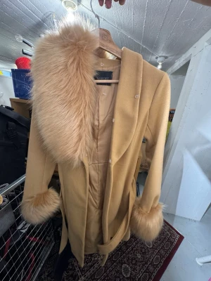 Beige kappa med fuskpälsdetaljer Garoff - Stilren beige kappa från Seaboff med stora fuskpälsdetaljer vid krage och ärmslut. Kappan har knytband i midjan och en klassisk, figurnära passform. Perfekt för dig som vill ha en elegant look med extra lyxig känsla. Finns att hämta i Gröndal 