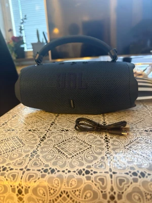 Blå JBL bärbar högtalare med handtag - Säljer en snygg blå JBL bärbar högtalare med robust handtag och tygklädd yta. Högtalaren har en cylinderformad design och tydlig JBL-logga framtill. USB-laddkabel medföljer. Perfekt för att ta med sig musiken överallt.