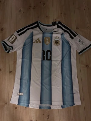 Argentina Messi matchtröja blåvit - Säljer en officiell Argentina matchtröja från Adidas med Messi och nummer 10 på ryggen. Tröjan har klassiska blå och vita ränder, svarta detaljer och guldiga loggor. Märken på ärmarna och broderade emblem på bröstet. Perfekt för fotbollsfans!
