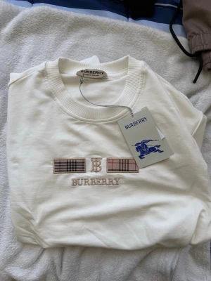 Vit sweatshirt från Burberry - Säljer en stilren vit sweatshirt från Burberry med klassiskt rutmönster och broderad logga på bröstet. Tröjan är gjord i 100% bomull och har rund halsringning. Perfekt för dig som gillar exklusiva och tidlösa plagg.