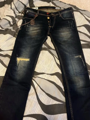 Mörkblå slitna jeans med kedja - Säljer ett par mörkblå jeans med slitningar och detaljer som kedja och patchade hål. Jeansen har kontrastsömmar, fem fickor och en cool streetwear-look. Perfekta för dig som gillar en edgy stil och vill sticka ut lite extra.