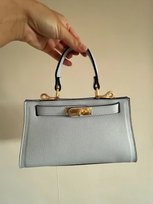 Ljusblå handväska med gulddetaljer - Säljer denna helt nya & oanvända väska från Don de Dieu Marque Made in Italy Genuine Leather. Köpt för 2253 kr (se bild) men säljer den för detta billiga pris eftersom jag vill bli av med den fort. Den är helt oanvänd, super snygg, babyblå/ljusblå, i äkta läder och har super fina guldiga detaljer. Denna väska är väldigt speciell och gjord i Italien, den liknar litegrann en annan populär väska ”mini Kelly”. Axelrem tillkommer. 