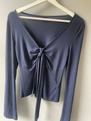 Mörkblå knytblus från Gina Tricot - Trendig mörkblå långärmad topp från Gina Tricot med v-ringning och snygg knytning framtill. Tunn och mjuk i materialet, perfekt till jeans eller kjol. Enkel men ändå unik detalj med knuten framtill som ger en cool look.