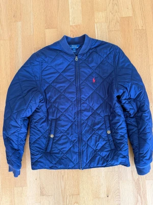 Marinblå quiltad jacka Polo Ralph Lauren - Snygg marinblå pufferjacka från Polo Ralph Lauren med quiltat mönster och röd broderad logga på bröstet. Jackan har ribbade muddar, dragkedja framtill och två fickor med tryckknappar. Perfekt höstjacka och riktigt stilren look. Junior Large 14-16 år
