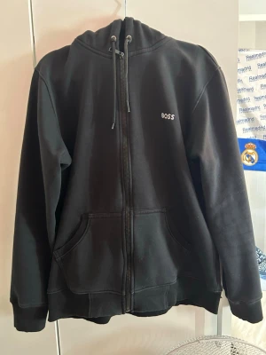 Svart zip hoodie från BOSS - Säljer en svart hoodie från BOSS med dragkedja framtill och två stora fickor. Tröjan har huva med dragsko och diskret BOSS-logga på bröstet. Tillverkad i mjukt sweatshirtmaterial som är perfekt för chill dagar.