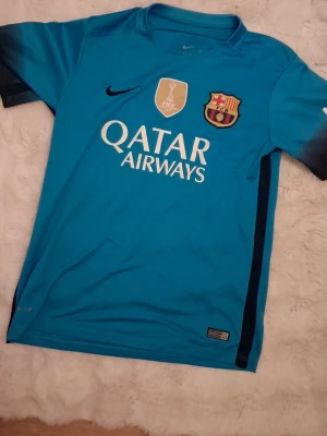 Blå FC Barcelona fotbollströja - Säljer en blå FC Barcelona fotbollströja med Qatar Airways-tryck på bröstet och klubbmärke samt FIFA-märke. Tröjan har korta ärmar och svart Nike-logga. Perfekt för dig som älskar fotboll och vill stötta Barca på match eller träning.