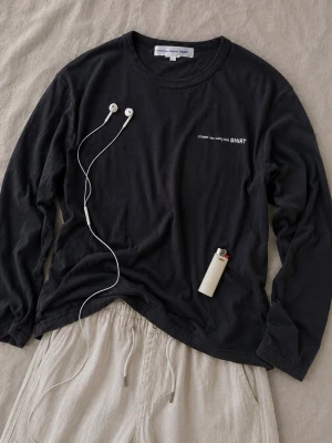 Svart långärmad topp Comme des Garçons - Svart långärmad topp från Comme des Garçons med rund hals och diskret vit text på bröstet. Tillverkad i mjuk bomull som känns skön mot huden. Perfekt för dig som gillar minimalistisk och clean stil. Passar M. 