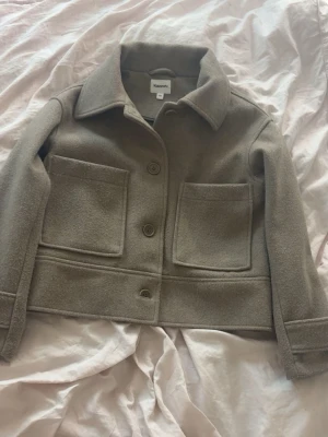 croppad jacka från Kappahl". - Stilren beige jacka från Kappahl i ullblandning. Jackan har stora framfickor, klassisk krage och knäppning med stora knappar framtill. Croppad modell med lång ärm och enkel, clean design som passar till många outfits.