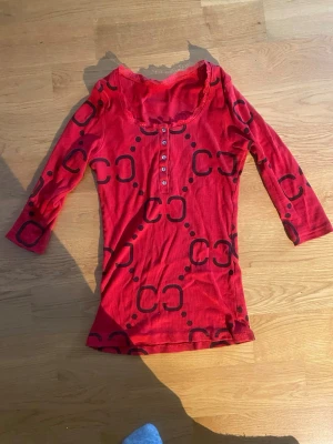 Röd topp med svart mönster och knappar - Röd topp med svart grafiskt mönster och trekvartsärm. Toppen har knappar framtill och en lätt rundad halsringning med spetsdetalj. Materialet känns mjukt och stretchigt, troligen bomull eller en bomullsblandning. Perfekt för dig som gillar färg och unika detaljer. Storlek står inte men jag tror de är mellan xs o s
