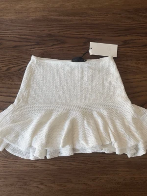 Vit volangkjol med struktur - Superfin vit kortkjol med volangkant och strukturerat tyg. Kjolen har en feminin och lekfull look, perfekt att styla med en enkel topp eller hoodie. Materialet känns mjukt och har en lätt stretch för extra komfort.
