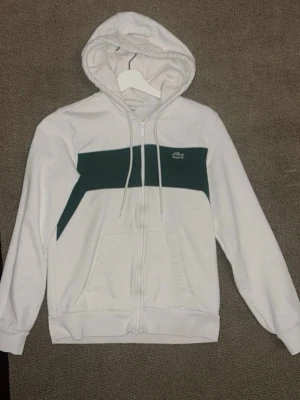 Vit Lacoste zip up - Snygg vit hoodie från Lacoste med dragkedja och en bred grön panel över bröstet. Klassisk krokodillogga på vänster sida. Utrustad med huva och dragsnören samt två fickor framtill. Perfekt för en avslappnad och sportig stil.