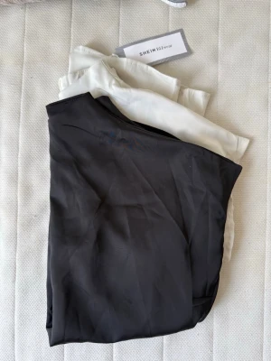 Två blusar från SHEIN i svart och vitt - Säljer två stilrena blusar från SHEIN, en i svart och en i vitt/beiget. Båda har korta ärmar och rak passform. Materialet (satin) känns lätt och mjukt, perfekt för en clean och enkel look. Passar till många olika outfits och är superlätta att matcha. Den svarta är använd några gånger men den vita är fortfarande helt ny med prislapp. 