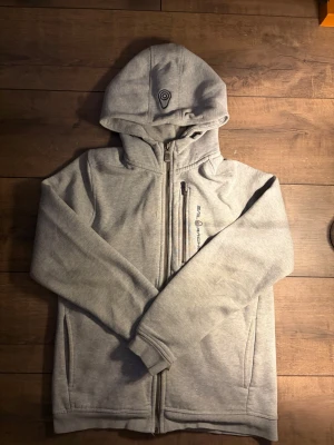 Sail racing kofta  - Snygg ljusgrå hoodie med dragkedja framtill och två fickor. Har en huva med tryckt detalj och diskret logga på bröstet. Tillverkad i mjukt sweatshirtmaterial som känns skönt mot huden. Perfekt för chill dagar eller när du vill ha en enkel och clean look.