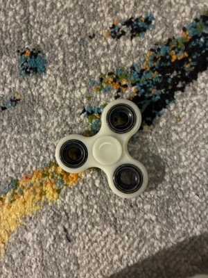Vit fidget spinner i plast - En klassisk vit fidget spinner med tre armar och svarta kullager. Tillverkad i plast och metall, perfekt för att hålla händerna sysselsatta eller för att slappna av. Enkel och smidig att ta med sig överallt. Den lyser över i mörkret efter att varit under en lampa.