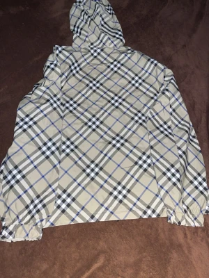 Beige rutig vindjacka med huva - Snygg beige vindjacka från Burberry med klassiskt rutmönster i svart, vitt och blått. Jackan har huva med dragsko, hel dragkedja framtill och elastiska muddar vid ärmsluten. Perfekt för dig som vill ha en stilren och trendig look.