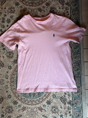 Ljusrosa t-shirt från Polo Ralph Lauren - Snygg ljusrosa t-shirt från Polo Ralph Lauren med klassisk passform och liten broderad logga på bröstet. Tillverkad i mjuk bomull som känns skön mot huden. Perfekt till jeans eller shorts för en clean och stilren look. Tyvärr kan den ha fläckar runt märket, men man märker nästan inte det, fin och bra för priset.