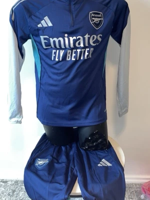 Arsenal träningsset blå adidas - Snyggt Arsenal träningsset i marinblått med vita detaljer. Setet består av en långärmad tröja med halv dragkedja och matchande byxor. Både tröjan och byxorna har Arsenal-logga och adidas-logga. Tillverkat i lätt och funktionellt material, perfekt för träning.