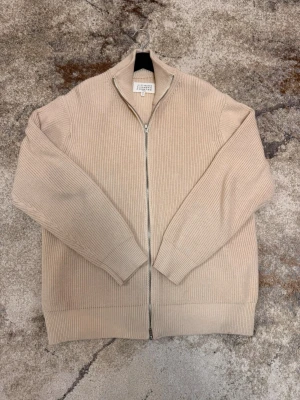 Beige stickad ziptröja Maison Margiela - Säljer en stilren beige stickad ziptröja från Maison Margiela. Tröjan har ribbstickad struktur, hög krage och dragkedja framtill. Perfekt för lager på lager och enkel att matcha med olika outfits.