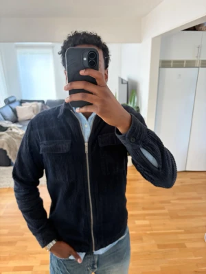 Mörkblå manchester overshirt - Säljer en stilren mörkblå overshirt i manchestertyg med dragkedja framtill och två bröstfickor med lock. Overshirten har klassisk krage och långa ärmar med knapp vid ärmslut. Perfekt att bära över en skjorta för en avslappnad look även stilig över en T-shirt med öppen look. Passar med olika typer av jeans. Liten defekt punkt i form av litet hål vid ärmen dock ingenting som bemärks när den bärs. Ordinarie pris 1199 kr från ASOS. 