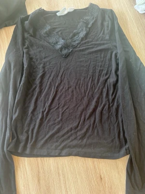 Svart långärmad topp med spets - Snygg svart långärmad topp från Gina Tricot med v-ringning dekorerad med spetsdetaljer. Perfekt att styla till jeans eller kjol för en trendig look. Mjuk och skön känsla i materialet.