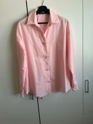 Ljusrosa skjorta med knyt - Säljer en ljusrosa skjorta med unika knytstängningar framtill. Skjortan har klassisk krage och långa ärmar, perfekt för dig som vill ha en stilren men ändå speciell look. Materialet känns mjukt och lätt, troligen bomull.