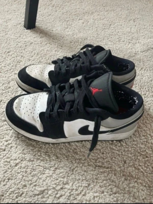 Svarta och vita Air Jordan 1 Low 38,5 - Jordan sneakers i svart och vitt med röda detaljer. Skorna har svarta skosnören och en vit sula. Jumpman-loggan syns på plösen och innersulan. Fint använt skick, lite noppriga bara