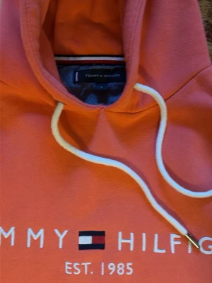 Korallfärgad hoodie Tommy Hilfiger - Snygg korallfärgad hoodie från Tommy Hilfiger med vit dragsko i huvan och stor magficka. Framtill står det 'Tommy Hilfiger EST. 1985' med den klassiska loggan broderad. Perfekt för en chill och trendig look.