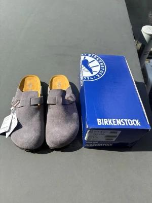 Grå Birkenstock Boston  - Säljer ett par snygga grå tofflor från Birkenstock i mjuk mocka. Modellen har en klassisk slip-in design med justerbart spänne och bekväm innersula i läder. Perfekta för chill dagar hemma eller när du vill ha något enkelt men stilrent på fötterna. Helt nya med box endast testade. Hör av er vid funderingar 