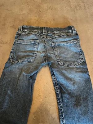 Blå slitna jeans med utsvängda ben - Säljer ett par blå jeans med utsvängda ben och klassisk femficksmodell. Jeansen har slitningar och tvättade detaljer för en avslappnad look. De har bälteshällor, dubbla knappar och dragkedja framtill. Perfekta för en casual stil.