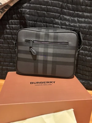  Burberry väska - Snygg svart axelväska från Burberry med klassiskt rutmönster i mörkgrå toner. Väskan har justerbar axelrem med präglad logga, dragkedja fram och bak samt detaljer i metall. Perfekt för dig som vill ha en stilren och trendig accessoar.