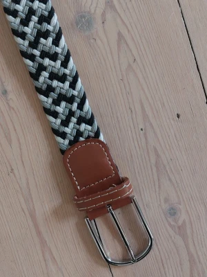 Randigt flätat bälte med läderdetaljer - Snyggt flätat bälte i svart, vitt och gråttmönster. Bältet har bruna läderdetaljer vid spännet och en klassisk metallspänne. 108 cm till spännet. 