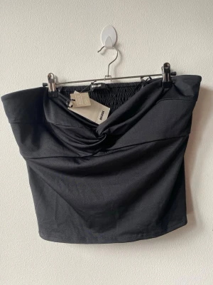 Svart axelbandslös topp från BikBok - Snygg svart axelbandslös topp från BikBok i storlek XL. Toppen har twistad detalj framtill och smockad rygg för extra komfort. Perfekt att styla med höga jeans eller kjol för en trendig look.