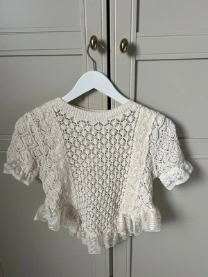 Vit virkad topp från Zara - Supersöt vit virkad topp från Zara med korta ärmar och volangkant nedtill. Toppen har ett detaljerat spetsmönster och rund halsringning. Perfekt för dig som gillar romantisk och vintage-inspirerad stil.