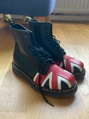 Dr Martens kängor Union Jack - Säljer ett par klassiska Dr Martens kängor i svart läder med Union Jack-flagga i rött, vitt och blått på tån. Grov sula med gul söm och högt skaft med AirWair-hälflik. Perfekta för dig som vill sticka ut med en ikonisk och edgy stil.