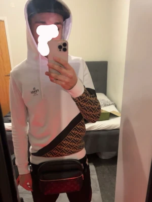 Vit hoodie från Fendi med logomönster - Säljer en snygg vit hoodie från Fendi med bruna partier och det klassiska Fendi-logomönstret på ena ärmen och sidan. Hoodien har en huva och svarta detaljer som ger en exklusiv vibe. Perfekt för dig som vill sticka ut med ett lyxigt streetwear-plagg.
