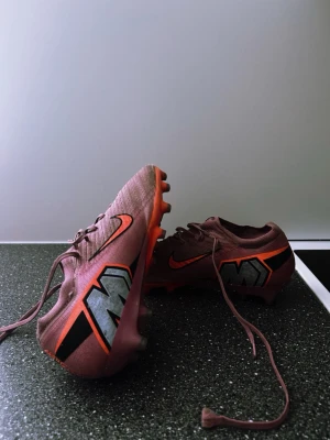 Rosa Nike Mercurial fotbollsskor - Säljer ett par rosa Nike Mercurial fotbollsskor med svarta och orange detaljer samt stor M-logga på sidan. Skorna har snörning och dobbar för spel på gräs. Tillverkade i syntetmaterial för bra grepp och känsla på planen.