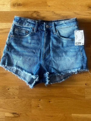 Blå high waist jeansshorts Hm - Säljer ett par blå high waist jeansshorts med råa, fransiga kanter och klassisk femficksdesign. Shortsen har knapp och dragkedja framtill och är perfekta för varma dagar. Från Hm