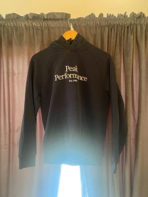 Svart Peak Performance hoodie - Säljer en svart hoodie från Peak Performance med vit logga och text 'Est. 1986' på bröstet. Hoodien har huva med dragsko och en stor magficka. Perfekt för dig som gillar en clean och klassisk stil.
