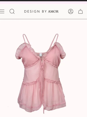 Rosa volang topp från Design by Amor - Supersöt rosa topp med tunna axelband och volangdetaljer från Design by Amor. Toppen har knyt framtill. Köpte för ca ett år sedan men den var för stor tyvärr. Aldrig använd. Köpte för 450kr.