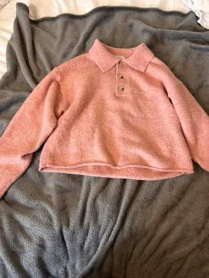 Rosa stickad tröja med krage - Mysig rosa stickad tröja med klassisk krage och tre knappar framtill. Tröjan har en boxy passform, långa ärmar och är tillverkad i ett mjukt, fluffigt material som känns skönt mot huden. Perfekt för kyliga dagar. är stor i storleken