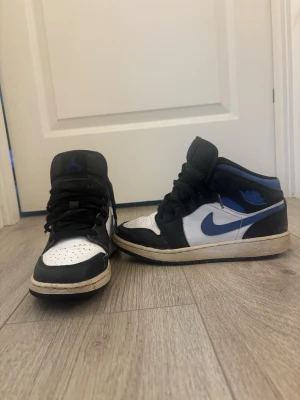 Nike Air Jordan 1 Mid svart/blå/vit - Säljer ett par Nike Air Jordan 1 Mid sneakers i svart, blått och vitt. Skorna har klassisk siluett med blå swoosh, svart läderpanel och vita partier. Perforerad tåbox och svart snörning. 