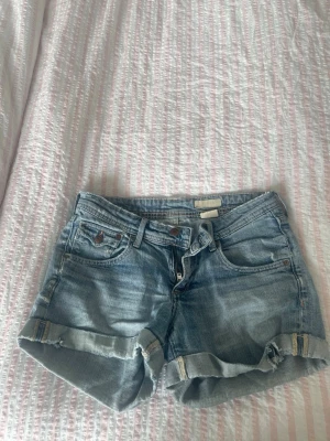 Ljusblå denimshorts med uppvik - Säljer ett par ljusblå denimshorts med uppvikta benslut och klassisk femficksdesign. Shortsen har bälteshällor, knappgylf och slitna detaljer för en avslappnad look. Perfekta för varma dagar och enkel att matcha med olika toppar.