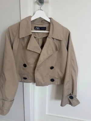 Kort beige trenchcoat från Zara - Stilren kort trenchcoat från Zara i beige bomullsmaterial. Jackan har dubbelknäppning med svarta knappar, bred krage och långa ärmar med knappdetalj vid ärmslut. Perfekt för en trendig och avslappnad look. En knapp fram som sönder bara👀