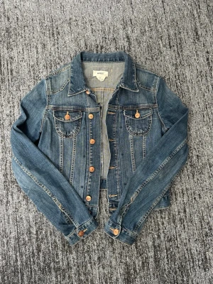 Klassisk blå jeansjacka från &jacket - Snygg blå jeansjacka från & jacket med klassisk krage, bröstfickor med lock och knappar i kopparfärg. Jackan har raka sömmar och en tidlös design som passar till det mesta. Tillverkad i slitstarkt jeansmaterial och har justerbara knappar i midjan.