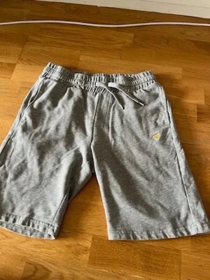 Grå mjukisshorts med snörning i nyskick - Snygga grå mjukisshorts i bomull med elastisk midja och snörning framtill. Shortsen har sidofickor och en bakficka samt en liten gul broderad logga på vänster ben. Perfekta för chill dagar eller träning.