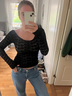Svart spetsig långärmad topp - Säljer en svart långärmad topp i spets med transparent mönster. Toppen har en rundad ringning och sitter tight mot kroppen. Perfekt att styla med jeans eller kjol för en trendig look.