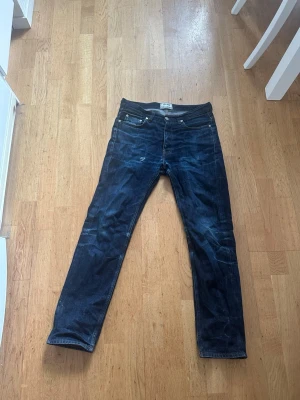 Raw denim acne jeans - Snygga mörkblå jeans med rak passform och klassisk femficksdesign. Jeansen har silverfärgade nitar och kontrastsömmar. Tillverkade i denim med en lätt tvättad look och subtila slitningar för en avslappnad stil. Väldigt bra skick men ett litet hål som jag själv tycker är snyggt