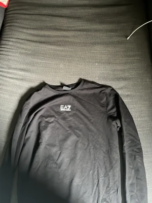 Svart långärmad tröja från EA7 - Snygg svart långärmad tröja från EA7 Emporio Armani med rund halsringning och vit logga på bröstet. Perfekt till jeans eller joggers för en clean och stilren look. Tillverkad i mjukt material som känns skönt mot huden.